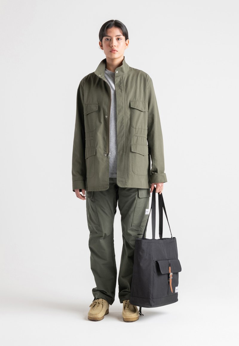 Maslinasto zelena utilitarna jakna s dva džepa na prsima, u kombinaciji s odgovarajućim cargo hlačama i crnom torbom s preklopom i kožnom trakom.