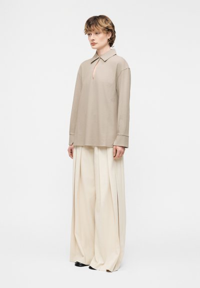 Άτομο με κοντά μαλλιά φοράει μπεζ μακρυμάνικο πουκάμισο με άνοιγμα στο λαιμό και φαρδιά κρεμ πλισέ παντελόνια, στέκεται μπροστά σε απλό φόντο.