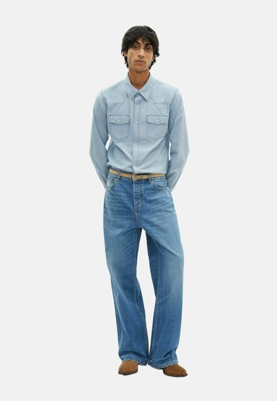 Camicia in denim azzurro chiaro con bottoni a pressione e due tasche sul petto, abbinata a jeans a gamba larga blu, completata da scarpe marroni.