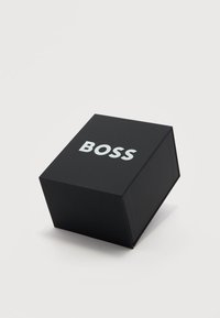 Μαύρο κουτί σε σχήμα κύβου με τη λέξη "BOSS" σε μεγάλα λευκά γράμματα στην κορυφή, τοποθετημένο σε απλό ανοιχτό φόντο.