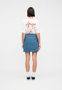Femme portant un t-shirt blanc avec le texte rouge "Amour" et deux lapins, une jupe en denim bleu, des chaussettes blanches et des baskets noires, debout de dos.