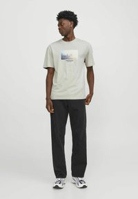 Camiseta de manga corta de color gris claro con un estampado gráfico en el centro, combinada con pantalones negros y zapatillas deportivas multicolor.