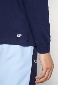 Námořnická košile s dlouhým rukávem a štítkem "ULTRA DRY"; spárovaná s světle modrými šortkami s zeleným krokodýlím logem a tmavým lemem.