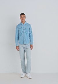 Lichtblauwe denim shirt met een knoopsluiting aan de voorkant, twee borstzakken en een klassieke kraag, gecombineerd met een lichte spijkerbroek en witte sneakers.