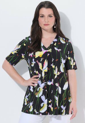 Femme aux longs cheveux foncés portant un haut court à manches courtes noir à motifs floraux et un pantalon blanc, debout avec une main sur la hanche, devant un fond blanc.