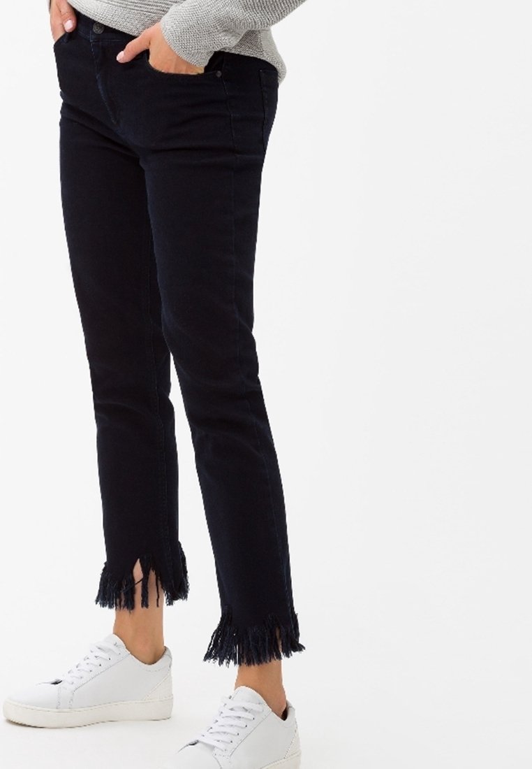 Jeans de mezclilla negra con un corte ajustado, dobladillo desgastado y longitud hasta el tobillo, combinados con zapatillas blancas. La textura parece resistente y ligeramente elástica.