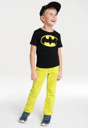 Black cotton T-shirt with yellow Batman logo, bright yellow pants, and blue athletic shoes. Child wearing a black cap, standing and smiling.