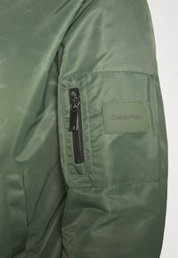Oliivinvihreä takki, jossa on sileä, kiiltävä pinta, vetoketjullinen tasku hihassa ja Calvin Kleinin logo-ompelu sivussa.
