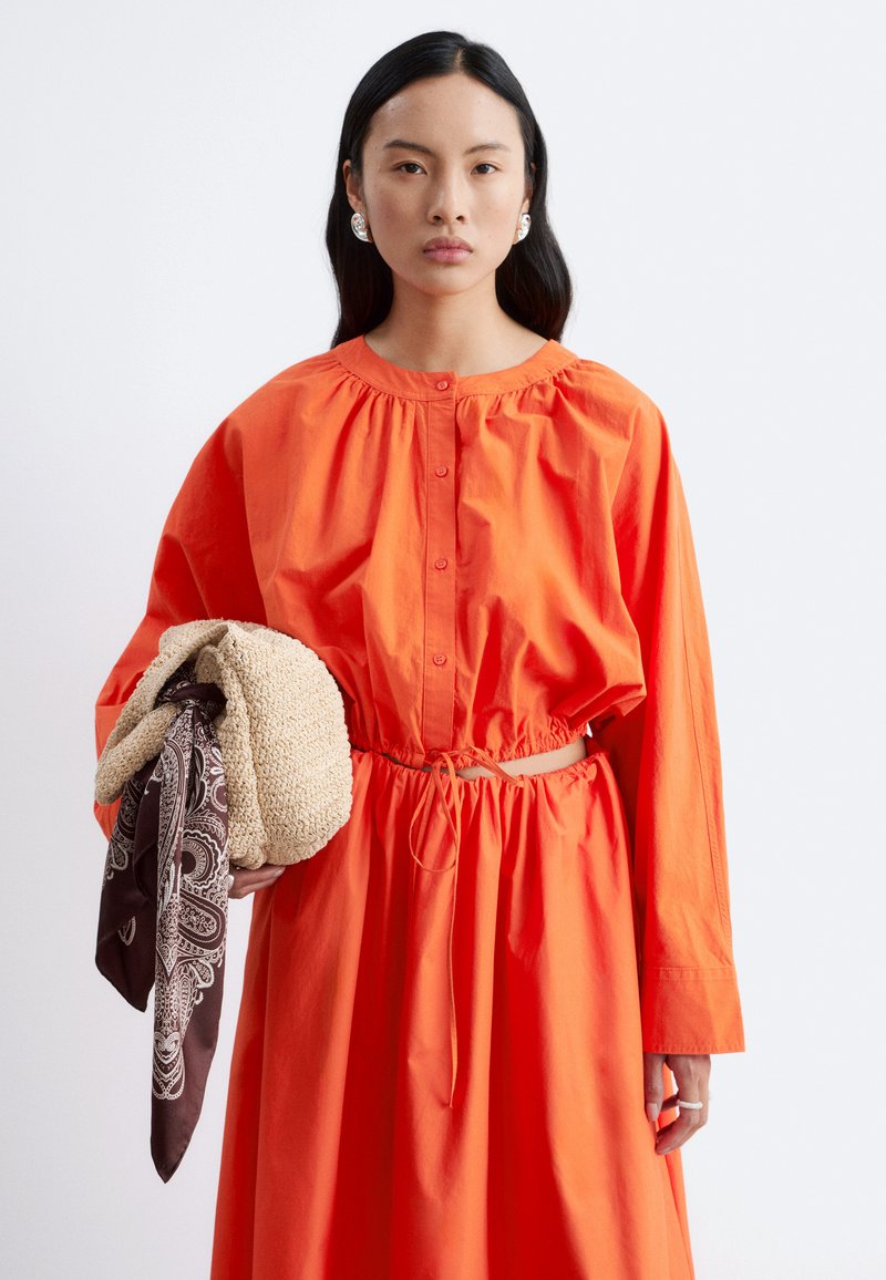 Femme aux longs cheveux foncés portant une robe ample orange vif, de grandes boucles d’oreilles argentées, tenant un sac tressé avec un foulard à motifs.