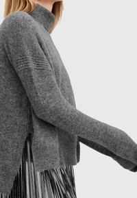 Femme portant un pull à col roulé gris doux avec des détails côtelés et une jupe plissée métallique sur un fond uni.