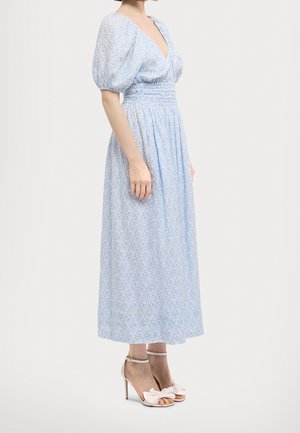 Frau trägt ein hellblaues Midikleid mit Blumenmuster, Puffärmeln, geraffter Taille und weißen Sandalen mit hohen Absätzen und Schleifen.