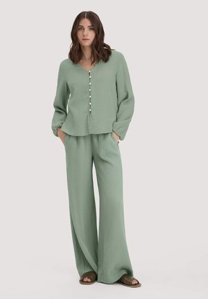 Frau steht mit den Händen in den Taschen, trägt eine locker sitzende, salbeigrüne, strukturierte, knopfbewehrte Bluse und passende, weit geschnittene Hosen mit offenen Sandalen.