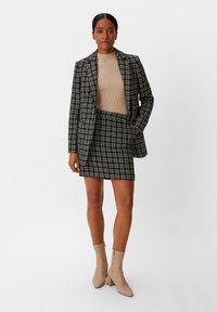 Schwarz-weißer karierter Blazer über einem beigen Top, kombiniert mit einem passenden Rock. Neutralfarbene Ankle Boots mit Blockabsatz.
