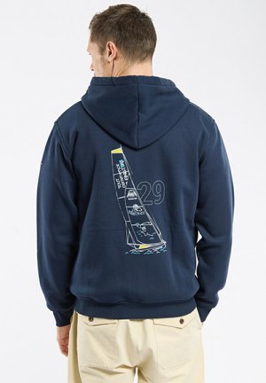 Mann trägt einen marineblauen Hoodie mit einem Segelboot-Grafik und der Nummer 29 auf der Rückseite, kombiniert mit beigefarbenen Hosen.