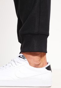 Ankelklädda i svarta joggers tillsammans med vita Nike-sneakers med svarta detaljer och vita snörningar mot en enkel bakgrund.