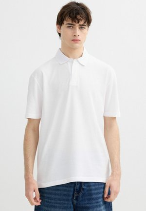 Joven con cabello oscuro que lleva una camiseta polo blanca lisa de manga corta y vaqueros azules, de pie frente a un fondo blanco.