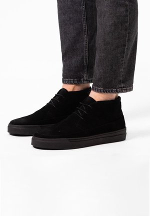Zwarte suède enkellaarzen met een gladde textuur, ronde neus en platte rubberen zool. Veters lopen over de bovenkant en hebben een minimalistisch ontwerp.