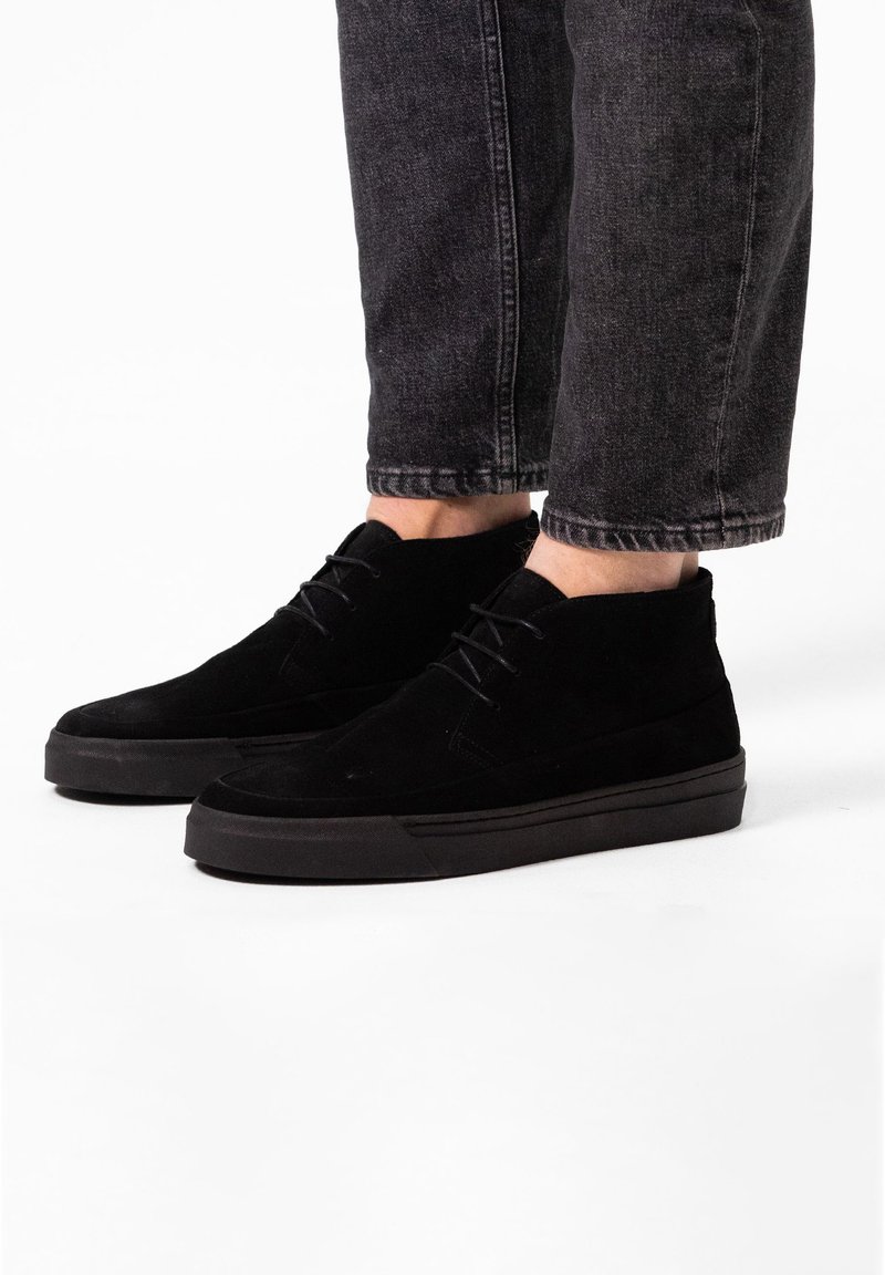 Stivaletti neri in suede con una texture liscia, punta tonda e suola piatta in gomma. I lacci si estendono sulla parte superiore, presentando un design minimale.