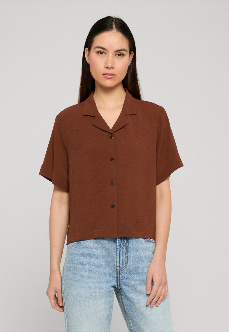 Femme aux longs cheveux foncés portant une chemise marron à manches courtes avec des boutons et un jean taille haute bleu clair, debout devant un fond uni.