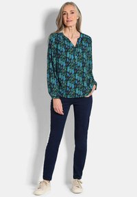 Grüne und blaue gemusterte Bluse mit V-Ausschnitt, langen Ärmeln und Knopfdetails. Kombiniert mit dunkelblauen Hosen und hellen Schuhen.