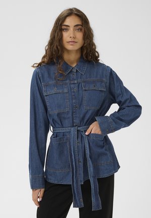 Sieviete, tērpusies zilā džinsa pogājā jakā ar divām krūšu kabatām, jostas siksnu, sasietu viduklī, un melnās biksēs, uz vienkārša balta fona.