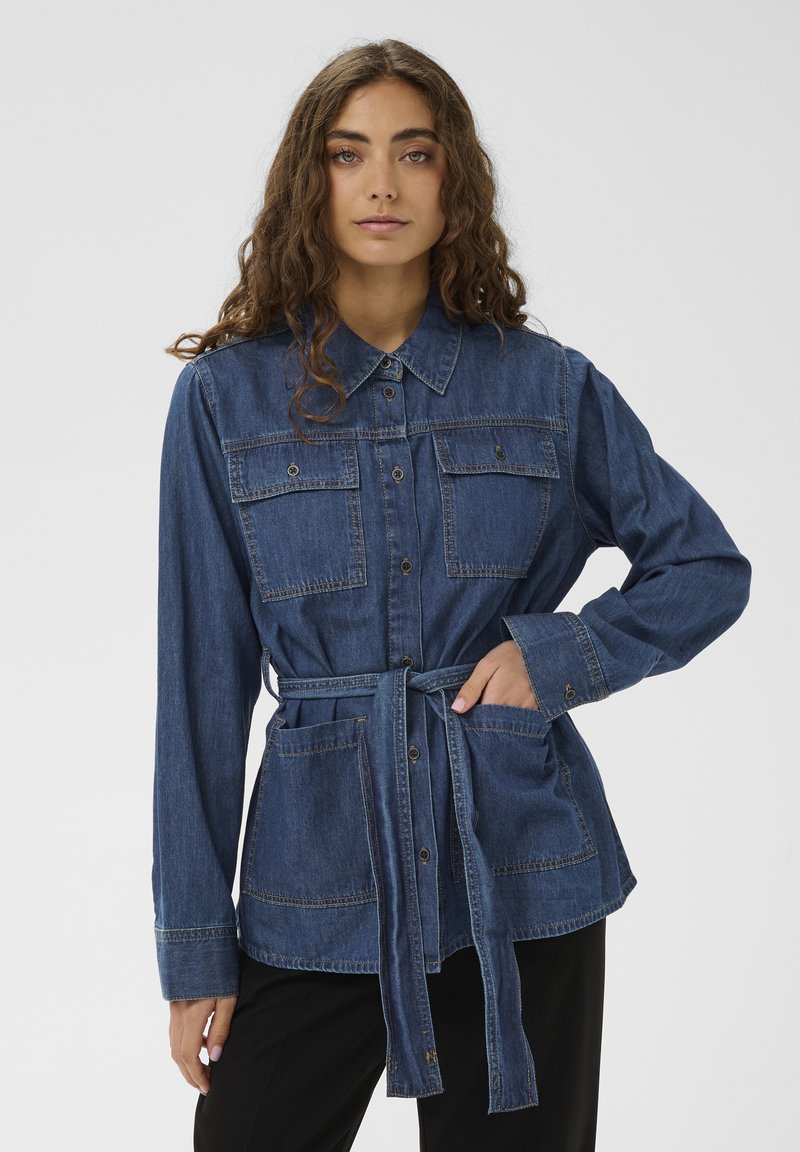 Kvinde iført en blå denimjakke med knapper, to brystlommer, et bindebælte i taljen og sorte bukser mod en ensfarvet hvid baggrund.