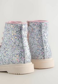 Bottines argentées brillantes avec une surface texturée, un col rembourré rose et une semelle blanche épaisse. Présente des paillettes en forme d'étoiles multicolores.