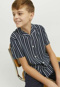 Camisa de manga corta color azul marino con rayas verticales blancas, de tela suave, cuello abierto y cierre frontal de botones, que lleva un niño sentado en una silla.