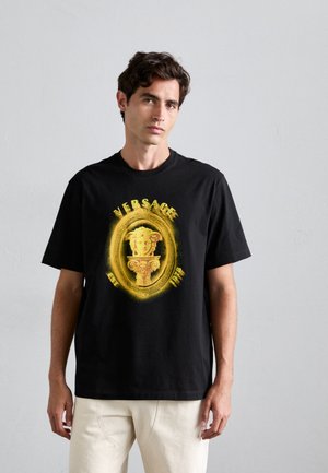 Mees, kes kannab musta Versace T-särki, millel on kollane Medusa pea ja sambakujundus, kombineerituna heledate pükstega, seisab sileda seina vastu.