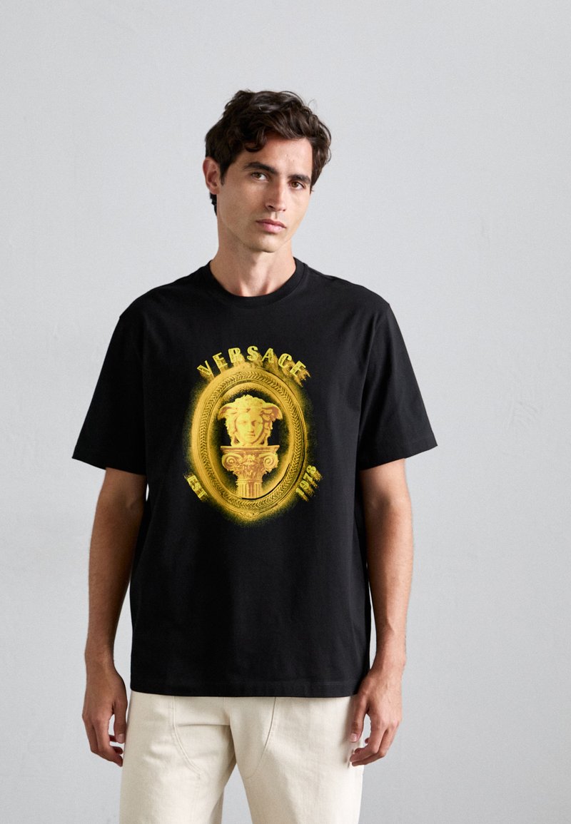 Man wearing black Versace t-shirt with yellow Medusa head and column design, paired with light-colored pants, standing against plain wall.