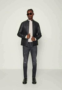 Jaqueta de imitação de pele preta com colarinho alto, combinada com uma blusa bege clara e calças de ganga escuras desgastadas. Botins pretos de tornozelo completam o visual.