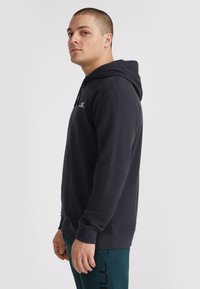 Haut à capuche noir en tissu doux, avec une poche kangourou, des poignets côtelés et un petit logo sur la poitrine. Coupe ajustée, design à enfiler.