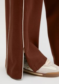 Καφέ παντελόνια με φαρδύ πόδι, λευκή ρίγα και πλαϊνό φερμουάρ, φορεμένα πάνω από μπεζ και λευκά αθλητικά παπούτσια.