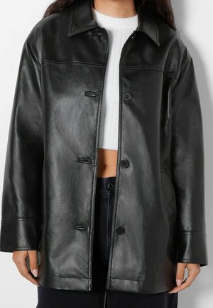 Person wearing a black leather button-up jacket over a white cropped sweater and black pants, hands resting by sides.