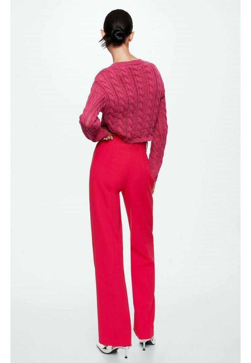 Femme aux cheveux foncés coiffés en chignon bas, portant un pull texturé rose et un pantalon large taille haute rose vif avec des talons blancs, vue de dos.