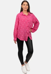 Camicia rosa con bottoni, decorata con motivi a forma di cuore bianchi, maniche lunghe e vestibilità rilassata, abbinata a leggings neri e sneakers bianche.