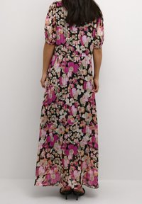 Femme portant une longue robe fleurie à manches bouffantes, debout de dos devant un fond uni.