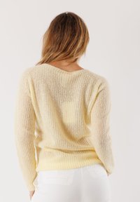 Donna con capelli lunghi che indossa un maglione lavorato a maglia color crema e pantaloni bianchi, voltata di spalle contro uno sfondo semplice.