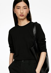 Schwarzes Langarmshirt mit Rundhalsausschnitt, kombiniert mit schwarzen Hosen. Eine glänzende schwarze Tragetasche wird auf der rechten Seite gehalten.