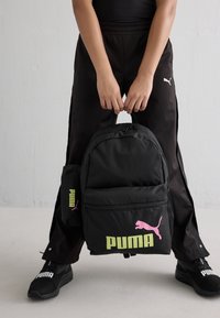 Juodas kuprinės su rožiniu Puma logotipu, turinti priekinį kišenę ir reguliuojamus diržus. Dėžutėje taip pat yra juodas pamušalinys su virvute.