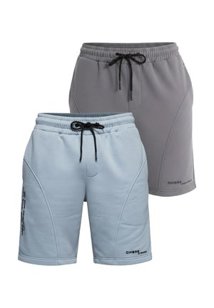 Homme debout portant un t-shirt jaune clair, un short bleu clair, des chaussettes blanches avec un logo noir et des baskets blanches sur un fond gris uni.