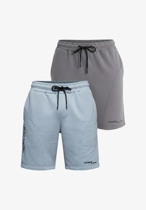 Homme debout portant un t-shirt jaune clair, un short bleu clair, des chaussettes blanches avec un logo noir et des baskets blanches sur un fond gris uni.
