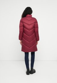 Giacca imbottita bordeaux con motivo a zigzag, colletto alto e maniche lunghe; indossata sopra jeans scuri e stivali neri.