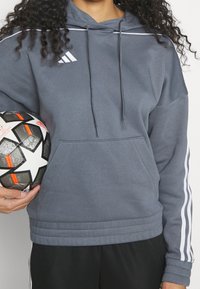 Un sweat à capuche gris avec une poche kangourou, un cordon de serrage pour la capuche, et des rayures blanches sur les manches. Tenant un ballon de football noir et blanc avec des accents orange.