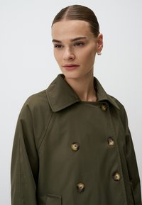Olivegreenowy dwurzędowy płaszcz trench z dużymi brązowymi guzikami i szerokim kołnierzem. Gładka tekstura, z bocznymi kieszeniami i przeszytymi detalami.