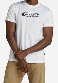 Camiseta blanca de manga corta con un logo impreso en negro "G-STAR" en el pecho. Fabricada en algodón con una textura suave.