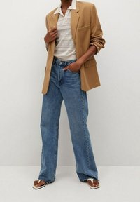 Beige Blazer über einem weißen strukturierten Stricktop, kombiniert mit hoch taillierten, weiten blauen Jeans und braunen Sandalen mit weißen Riemen.