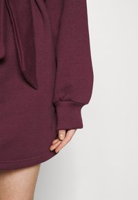 Bordeaux Sweatshirtkleid mit lockerer Passform, gerippten Bündchen und Details mit gebundenem Taillenband. Weicher Stoff mit glatter Oberfläche.