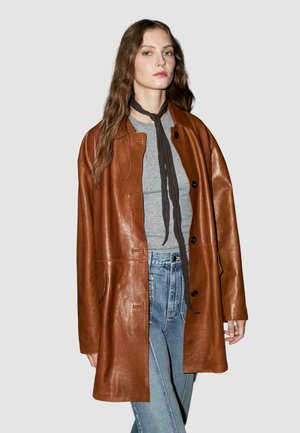 Manteau en cuir marron avec une coupe ample, doté d'une fermeture à boutons, de grandes poches et d'une texture lisse et brillante. Porté sur un haut gris et un jean bleu.