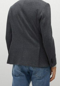 Uomo che indossa un blazer a spina di pesce grigio scuro e jeans blu, mostrato di spalle con la mano destra rilassata al fianco.